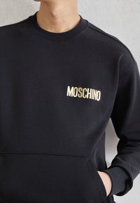 Černá mikina vyrobená z bavlněné směsi, se zlatým logem "MOSCHINO" na levém prsu a přední kapsou. Hladká textura, design s kulatým výstřihem.