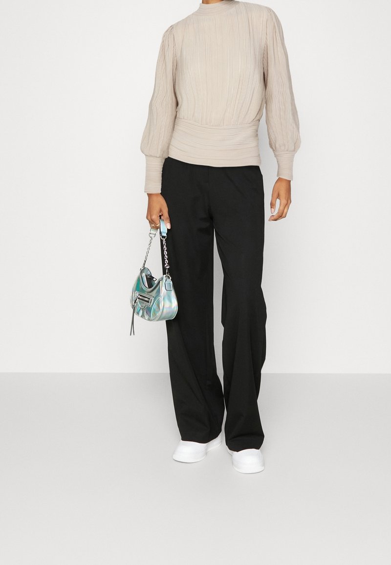 Personne portant un haut beige texturé à manches longues, un pantalon noir à jambes larges, des baskets blanches, tenant un petit sac à main métallique par la chaîne.