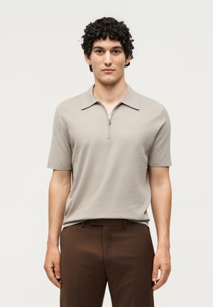 Homme aux cheveux bouclés foncés portant un polo beige à manches courtes avec une fermeture éclair et un pantalon marron, debout devant un fond clair uni.