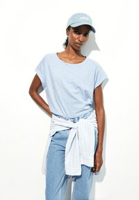 Lichtblauw gestreept T-shirt met korte mouwen, gecombineerd met lichtblauwe jeans en een gebonden wit overhemd. Pet in zachtblauw.