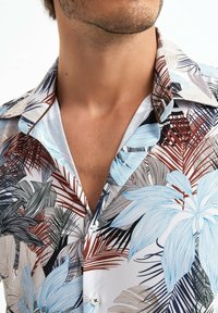 Chemise à manches courtes en tissu léger avec un motif floral bleu, marron et noir sur fond blanc.
