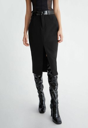 Kvinde iført en sort pencilnederdel med høj talje og frontslids, sort lakbælte og skinnende sorte knæhøje støvler, stående mod en ensartet baggrund.