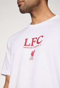 Λευκό βαμβακερό μπλουζάκι με κόκκινο κείμενο "LFC ANFIELD RD, L4" και κόκκινη σφραγίδα της Liverpool FC, με στρογγυλή λαιμόκοψη και κοντά μανίκια.