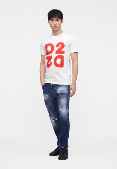 Jeune homme portant un t-shirt blanc avec de grands chiffres rouges et un jean bleu déchiré, debout avec une main dans la poche et des baskets noires.
