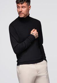 Czarny sweter w serek z długimi rękawami, prążkowanymi mankietami i gładką fakturą, zestawiony z jasnymi spodniami. Mężczyzna w swetrze pozuje.