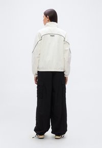 Λευκό ελαφρύ μπουφάν με μαύρες λεπτομέρειες, που διαθέτει ψηλό γιακά και ελαστικές μανσέτες, συνδυασμένο με μαύρο παντελόνι cargo φαρδιάς γραμμής.