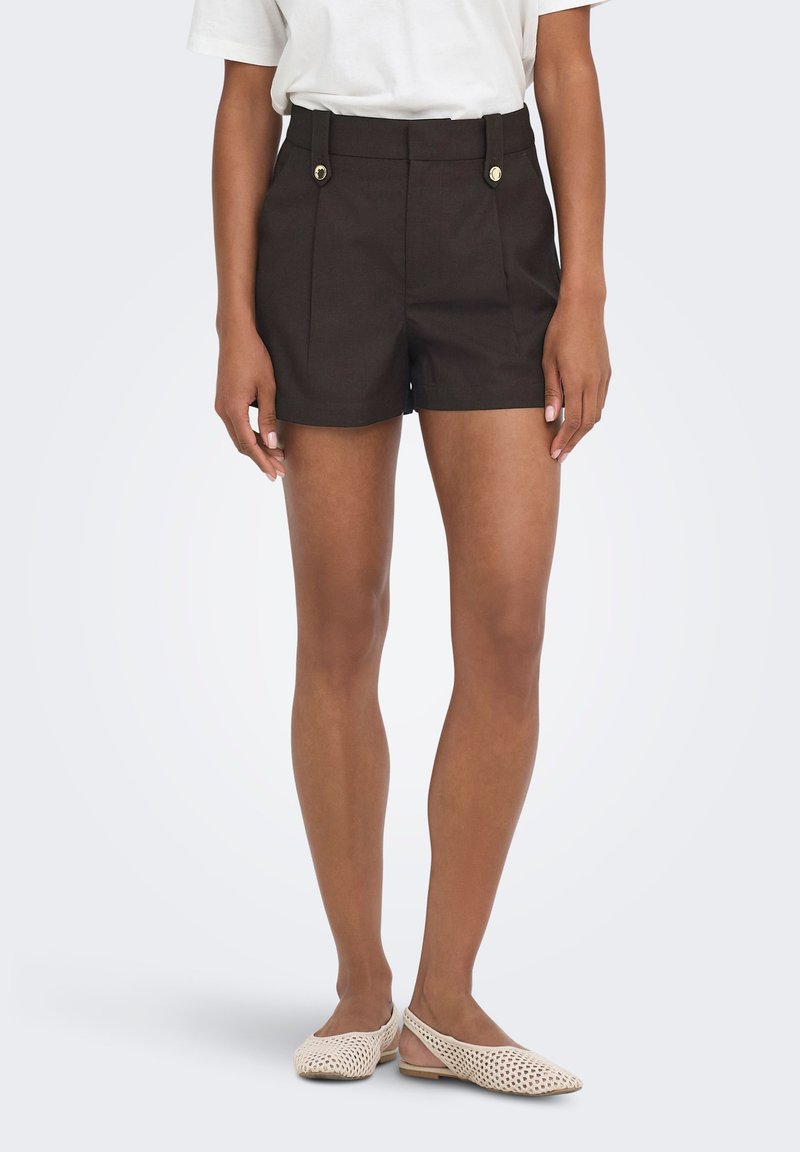 Femme portant un short noir taille haute avec des boutons dorés, un haut blanc et des ballerines beiges tissées à bride arrière, debout devant un fond uni.