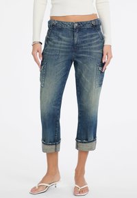 Blaue Denim-Cargohosen mit umgeschlagenen Säumen, Fronttaschen und seitlichen aufgesetzten Taschen. Der Stoff zeigt eine ausgewaschene Optik und eine strukturierte Oberfläche.