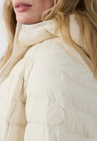 Roomkleurig donzen jack met een hoge kraag, horizontale stiksels en een JOTT-logo op de mouw. Zachte, gewatteerde textuur.