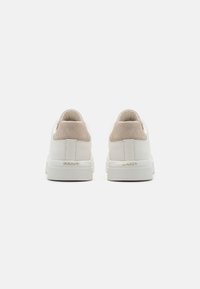 Paire de baskets blanches avec des contreforts en daim beige et le logo "GANT" en or à l'arrière des semelles en caoutchouc blanc, vues de dos.