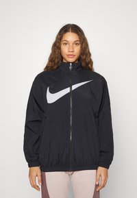 Mustat Nike-tuulenpitäjä, jossa on korkea kaulus, täysipitkä vetoketju ja suuri valkoinen logo. Varustettu jousto-ompeleet hihoissa ja kevyellä, teksturoidulla kankaalla.