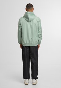 Světle zelená bunda s kapucí a elastickým pasem, v kombinaci s černými kalhotami a bílými teniskami, pohled zezadu.