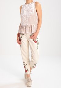 Top peplum in velluto rosa chiaro abbinato a pantaloni crema con ricami floreali rossi. Modella in sneakers beige, in piedi contro uno sfondo neutro.