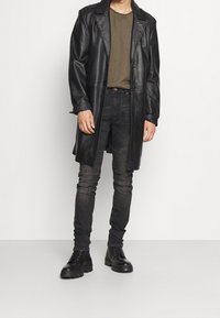 Hombre con abrigo de cuero negro, camiseta verde oliva, jeans negros texturizados y botas negras de cordones de pie contra un fondo blanco.