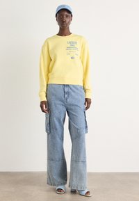 Felpa gialla con logo Lacoste blu, abbinata a pantaloni in denim a gamba larga con tasche cargo e finitura testurizzata. Cappellino blu e scarpe aperte.
