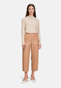 Beige Langarmoberteil mit hohem Kragen, kombiniert mit sandfarbenen, plissierten, weit geschnittenen Kunstlederhosen. Weiße spitze Ballerinas vervollständigen den Look.