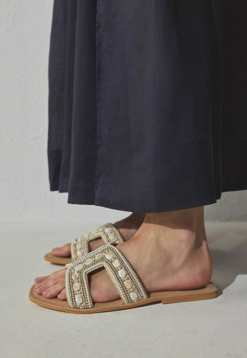 Füße in beigen flachen Sandalen mit Perlen- und Perlenverzierungen, kombiniert mit einem dunklen Rock oder einer Hose bis zum Knöchel.