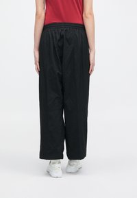 Άτομο που στέκεται γυρισμένο, φορώντας φαρδιά μαύρα παντελόνια, κόκκινο μπλουζάκι, λευκά αθλητικά παπούτσια, με τα χέρια χαλαρά πλάι στο σώμα, σε απλό ανοιχτό φόντο.