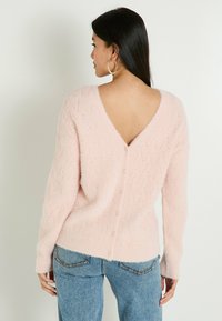 Pull en tricot rose clair avec un dos en V, un motif texturé, des manches longues, et des détails de boutons le long de la colonne vertébrale, assorti à un jean bleu.