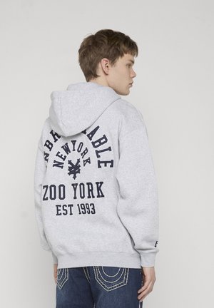 Jeune porte un sweat à capuche gris clair avec un texte bleu foncé "UNBREAKABLE NEW YORK ZOO YORK EST 1993" au dos, assorti à un jean bleu.