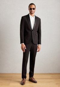 Traje negro con solapas de satén, camisa blanca con botones y zapatos de vestir marrones. El modelo lleva gafas de sol oscuras y está de pie sobre un suelo de madera.
