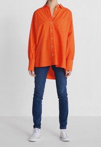 Chemise orange à manches longues avec des boutons, dotée d'un col et d'une poche poitrine. Le tissu semble lisse ; coupe ample avec un ourlet incurvé.