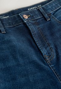 Sötétkék, hordó szabású farmernadrág. Elöl gombos záródással és kontrasztos varrással rendelkezik. Simább textúra, finom elhalványulásokkal.