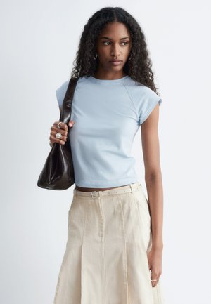 Jeune femme aux cheveux bouclés portant un haut bleu clair à manches courtes, une jupe beige, portant un sac noir à l’épaule, regardant sur le côté.