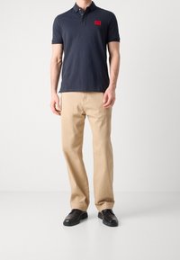 Granatowa koszulka polo z czerwoną logo, krótki rękaw i zapięcie na trzy guziki, połączona z beżowymi spodniami straight-leg i czarnymi butami.