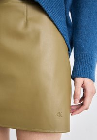 Kaki skinnminiskjørt med en glatt tekstur, med en subtil preget logo. Kombinert med en blå strikkegenser.