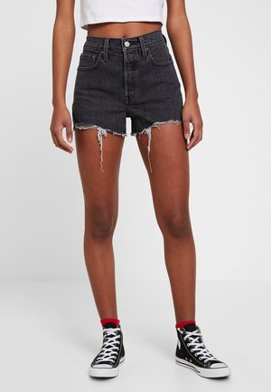 Personne debout portant un short en jean noir taille haute avec des ourlets effilochés, un top court blanc, des baskets hautes noires et des chaussettes rouges.