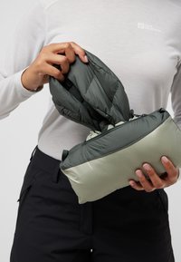 Giacca a vento verde scuro riposta in una borsa rettangolare verde chiaro. Il materiale sembra liscio e leggero con chiusura a zip.