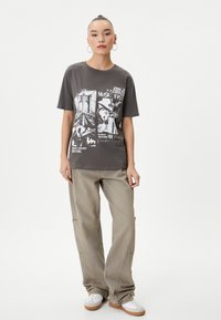 Grå grafisk t-shirt med hvidt print, der viser en guitar og "2002 MUSIC FEST." Bæret med lyse beige løstsiddende cargo-bukser og hvide sneakers.