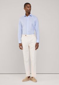 Camisa de manga larga azul claro con cuello puntiagudo, pantalones crema a medida y zapatos tipo mocasín beige con acabado texturizado.
