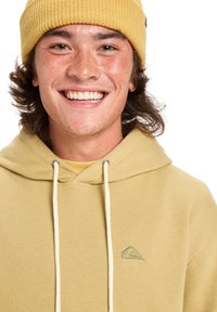 Gorro de punto amarillo y sudadera a juego con cordón, que presenta un pequeño logo verde en el pecho. Textura suave, ajuste casual.