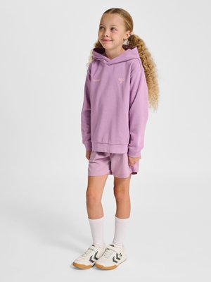 Mädchen mit lockigem Haar, das einen fliederfarbenen Hoodie, passende Shorts, weiße Socken und weiße Sneaker mit schwarzen Chevron-Logos trägt.