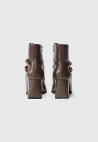 Par de botas de cuero marrón oscuro hasta el tobillo con tacones de bloque y dos hebillas laterales, mostradas desde atrás sobre un fondo blanco liso.