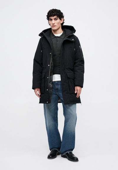 Ilgas juodas parka su kepure, turintis užtraukiamas šonines kišenes, priekinę užsegimą su sagomis ir siuvinėtą pamušalą, derinamas su plačių kojų mėlynomis džinsomis.