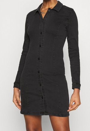 Robe en jean noire à manches longues avec col et boutons portée par une personne, présentant des coutures ajustées et une longueur mi-cuisse.