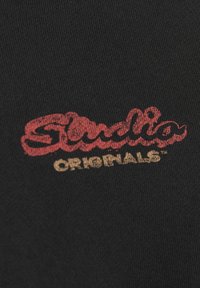 T-shirt nera in cotone con logo stampato rosso e oro contenente il testo “Studio Originals” in un carattere in corsivo vintage.