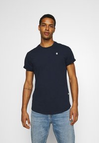 Himmelblaues Baumwoll-T-Shirt mit Rundhalsausschnitt und kurzen Ärmeln. Verfügt über ein kleines Logo auf der Brust und ein Etikett am Saum. Getragen mit hellblauen Jeans.