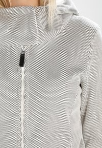 Chaqueta de cremallera gris claro con un patrón texturizado de panal, con un alto cuello y un tirador de cremallera negro para un cierre seguro.