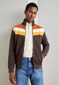 Braune Zip-Jacke mit horizontalen Streifen in Orange, Weiß und Gelb auf der oberen Brust. Aus weichem Stoff gefertigt, mit hohem Kragen.