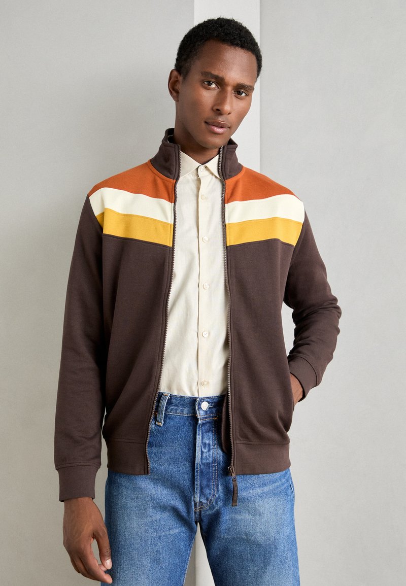 Braune Zip-Jacke mit horizontalen Streifen in Orange, Weiß und Gelb auf der oberen Brust. Aus weichem Stoff gefertigt, mit hohem Kragen.
