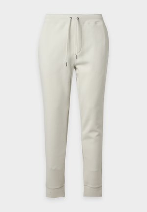 Giovane uomo che indossa una t-shirt blu navy con stampe di uccelli, pantaloni jogger bianco sporco e sneakers bianche, in piedi con una mano in tasca.