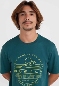 Teal bomulds t-shirt med rund hals, gul grafisk design med bølger og sol, mærkenavn "O'Neill" og tekst trykt på forsiden.