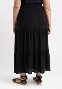 Gonna maxi a strati nera con fascia elastica, realizzata in tessuto leggero. Indossata con espadrillas nere caratterizzate da una trama intrecciata e lacci in raso.