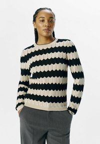 Gestrickter Pullover mit einem Zickzackmuster in Schwarz und Beige. Verfügt über lange Ärmel, einen gerippten Halsausschnitt und ein strukturiertes Gewebe. Kombiniert mit grauen Hosen.