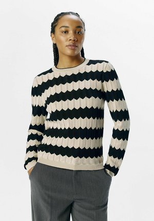 Strikket sweater med et zigzag mønster i sort og beige. Har lange ærmer, ribbet halsudskæring og tekstureret stof. Bæres med grå bukser.