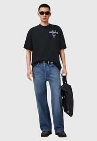 Sort oversized bomulds t-shirt med grafisk tryk, blå lige jeans, sort bælte og solbriller, holder en sort taske.